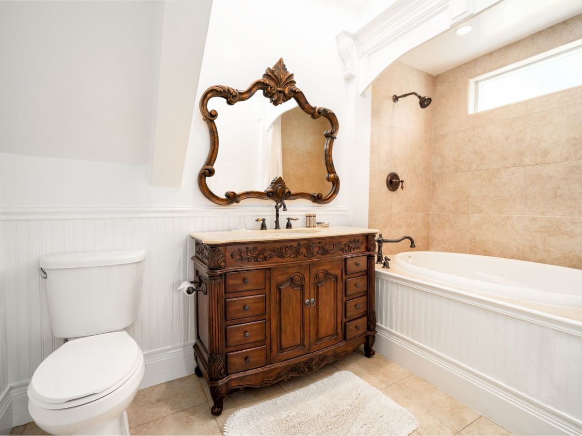 A classic bathroom with a wooden ornate vanity, an elegant curved mirror, a white toilet, a built-in bathtub, and beige tiled walls.