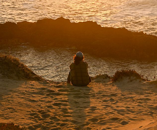 A lone person sits on the rocky shore as the sun sets the ocean in gold, peaceful and reflective.