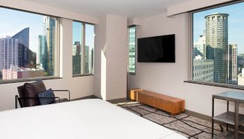 A modern hotel room with a large bed, a TV on the wall, floor-to-ceiling windows, and a view of city skyscrapers.