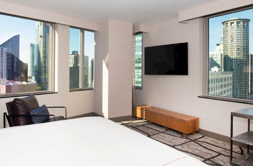 A modern hotel room with a large bed, a TV on the wall, floor-to-ceiling windows, and a view of city skyscrapers.