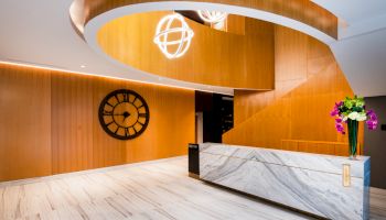 A modern interior with a marble reception desk, a large clock on a wooden wall, and geometric light fixtures hanging from the ceiling.