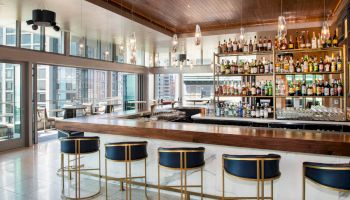 A modern bar with a wooden counter, four bar stools, shelves of various bottles, hanging lights, and large windows offering a city view ends the sentence.