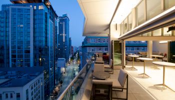 The image shows a modern cityscape with high-rise buildings and a rooftop patio featuring tables, chairs, and an indoor seating area.