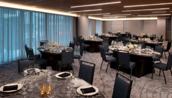 The image shows an elegant banquet hall with round tables, decorated with black napkins, glasses, and floral centerpieces, illuminated by ambient lighting.