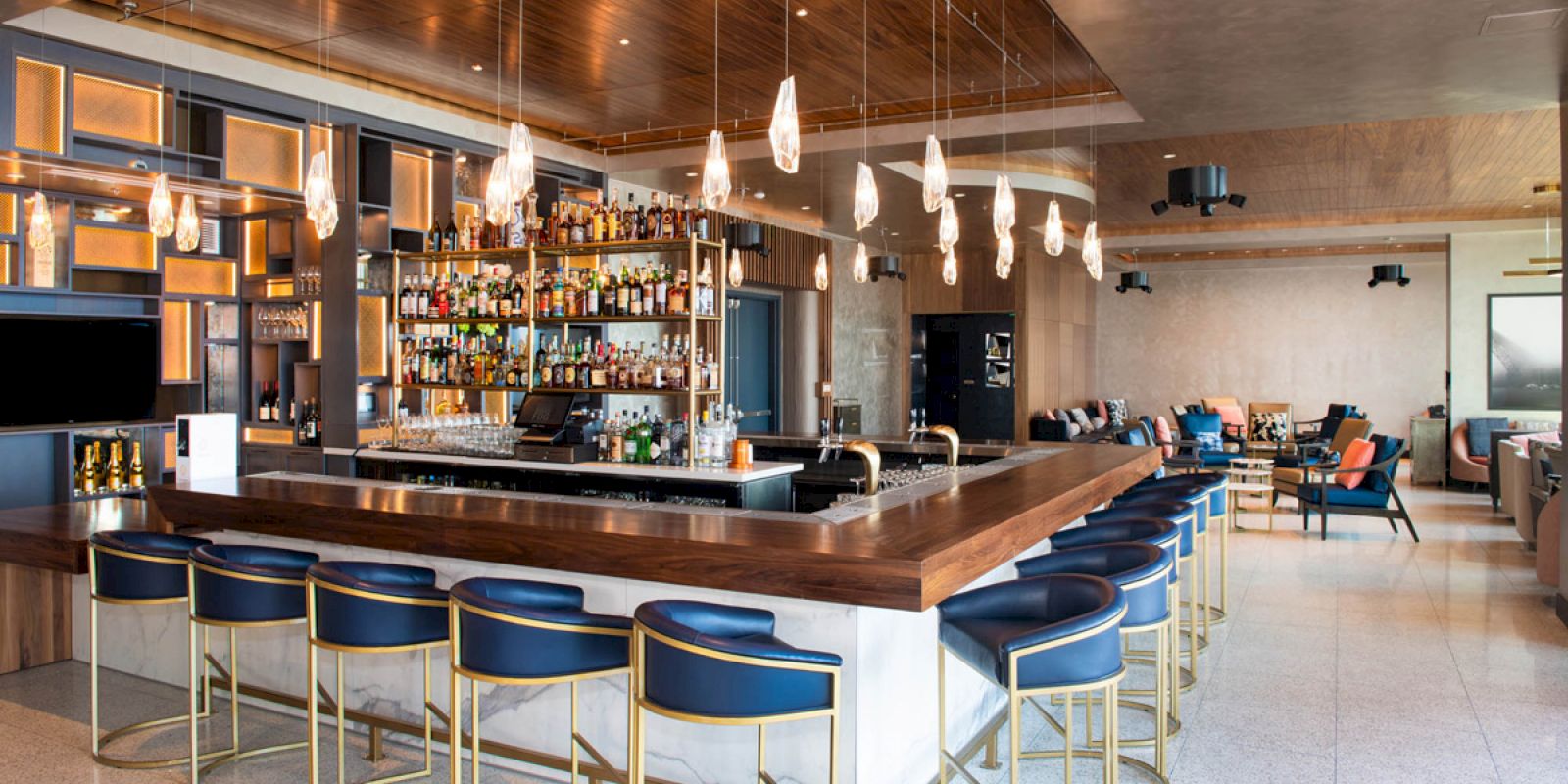 The image shows a modern bar with blue stools and a well-stocked bar counter, surrounded by elegant lighting and seating areas in contemporary decor.