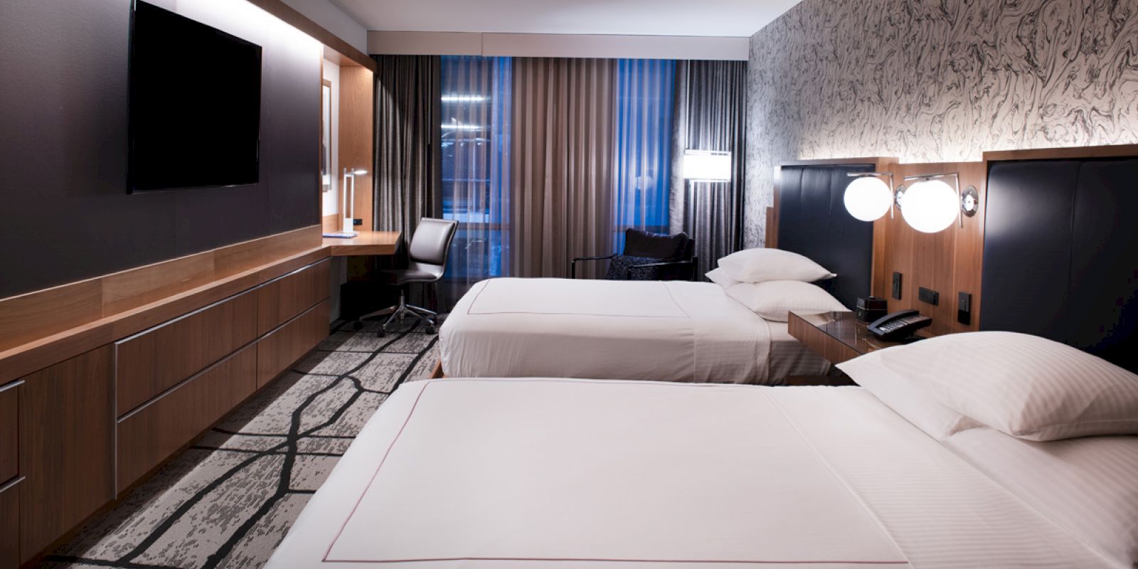 A modern hotel room with two beds, a large flat-screen TV, a desk with a chair, wall lamps, and a window with curtains.