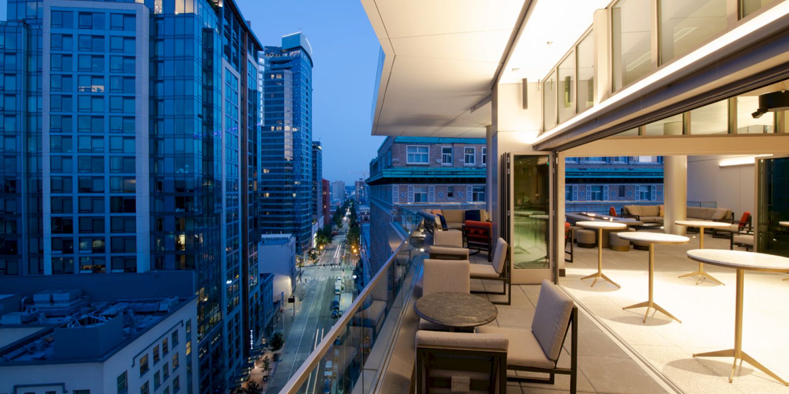 This image shows a modern cityscape with tall buildings, a street view, and a balcony featuring tables, chairs, and an outdoor seating area.
