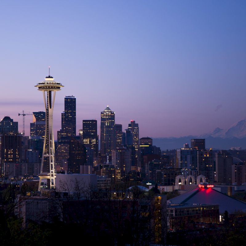 The image depicts a twilight view of Seattle's skyline, featuring the iconic Space Needle and Mount Rainier in the background.