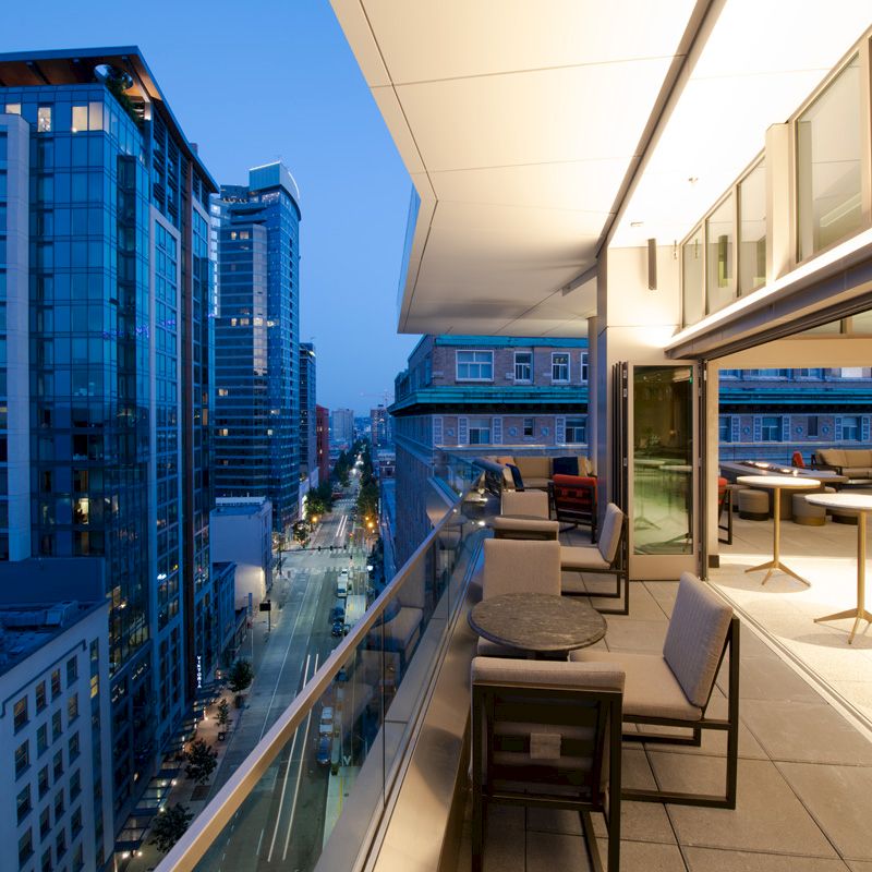 The image shows a modern cityscape with tall buildings and a rooftop patio with furniture, overlooking a street at dusk.