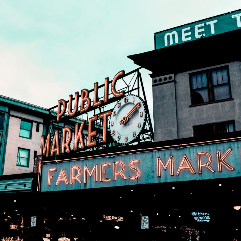 A neon sign at a public farmers market with a clock is mounted on a building. Other buildings are visible in the background.