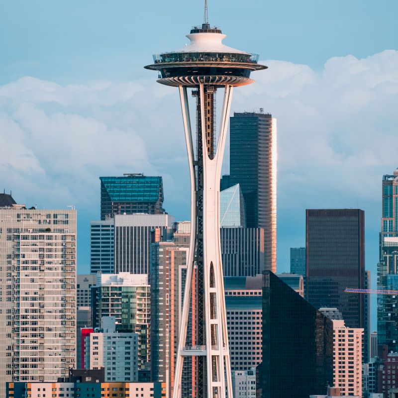 The image shows the Space Needle against a backdrop of tall buildings in a cityscape. The sky is clear, and the overall scene is bright and vibrant.