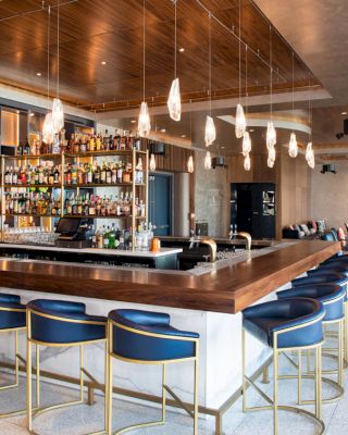 A modern bar with a wooden counter, blue and gold bar stools, hanging pendant lights, a well-stocked liquor shelf, and a lounge area in the background.
