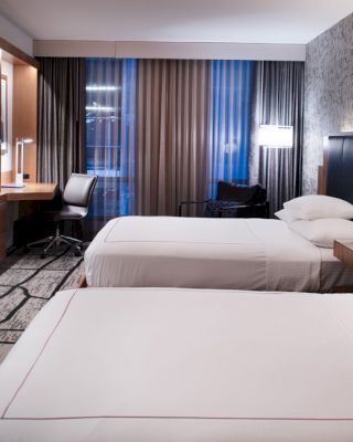 A modern hotel room with two neatly made beds, a wall-mounted TV, a desk, chair, bedside lamps, and contemporary decor.