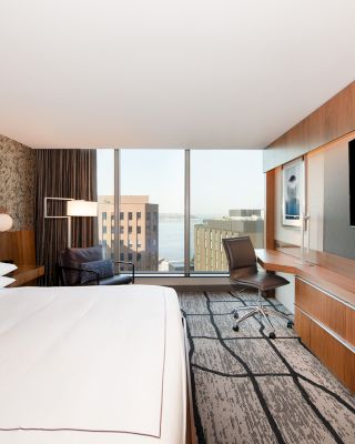 A modern hotel room with a large bed, a desk, a chair, a flat-screen TV, and a large window offering a cityscape view.