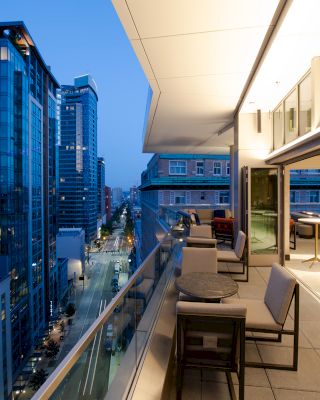 An outdoor terrace with chairs and tables is looking over a city skyline with modern buildings during dusk hours.