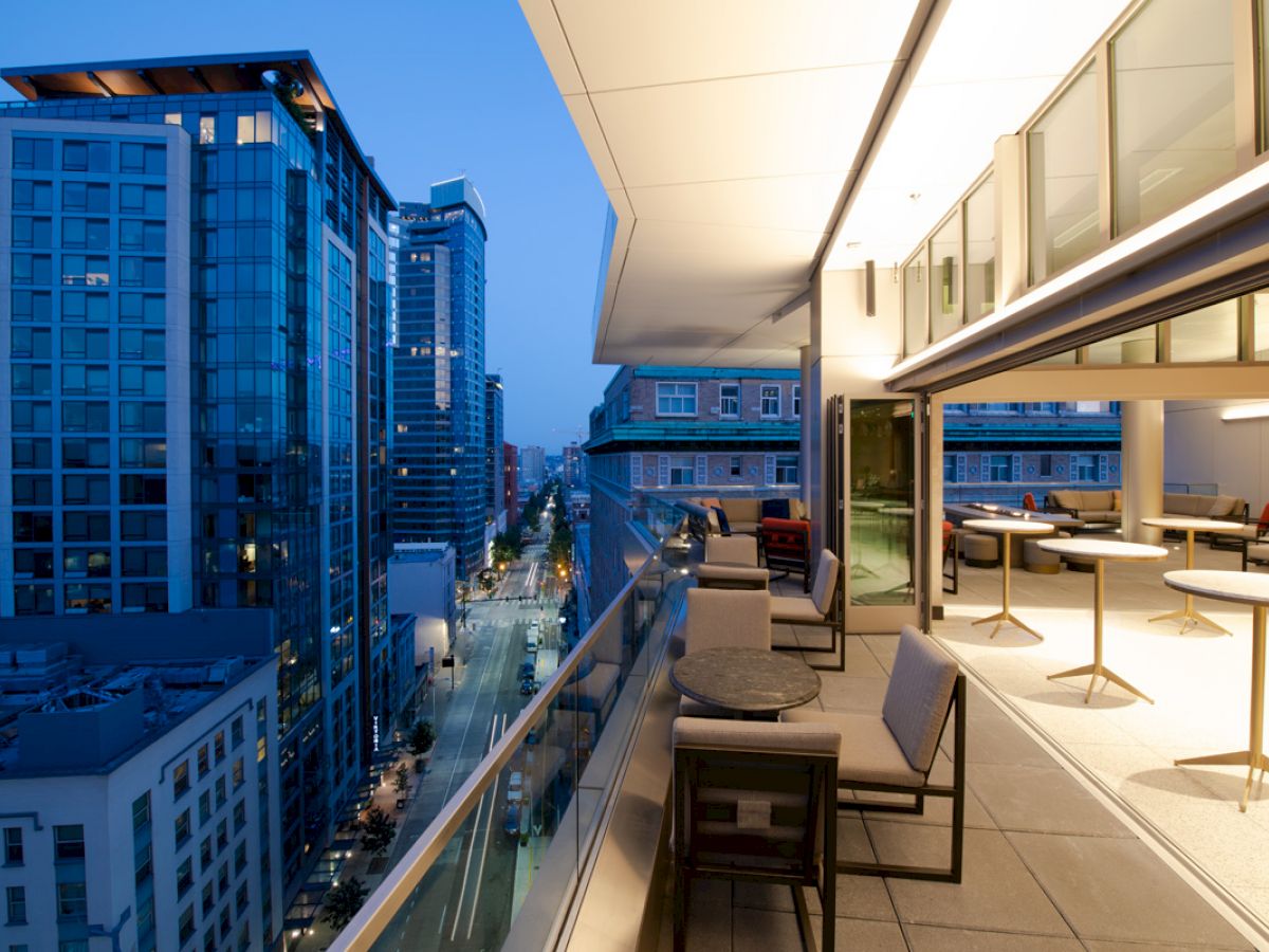 An evening cityscape view from a modern balcony with tables and chairs, overlooking tall buildings and a street illuminated by lights.