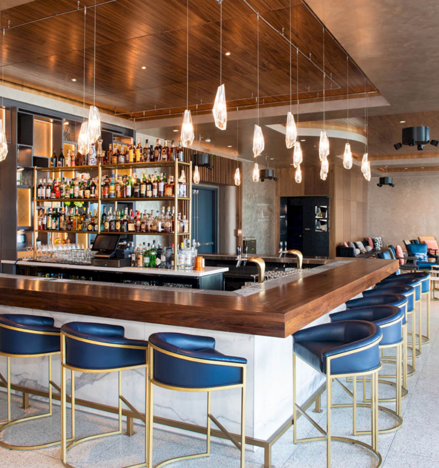 The image shows a modern bar with stylish blue and gold bar stools, a fully stocked bar, and hanging pendant lights in a well-designed, spacious interior.
