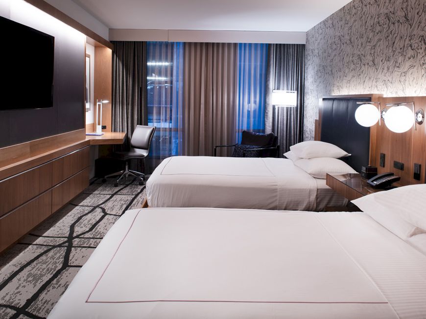 A modern hotel room features two neatly made beds, a flat-screen TV, a desk with a chair, wall art, and soft lighting.