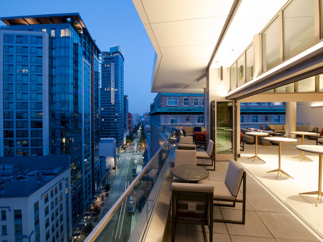 A modern balcony overlooks a cityscape with tall buildings at dusk. The stylish outdoor area has chairs, tables, and opens into an indoor space.