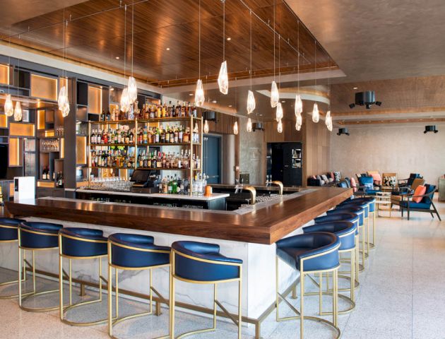 A modern bar with a wooden countertop, blue chairs, hanging lights, and a well-stocked liquor shelf in an elegantly decorated space.
