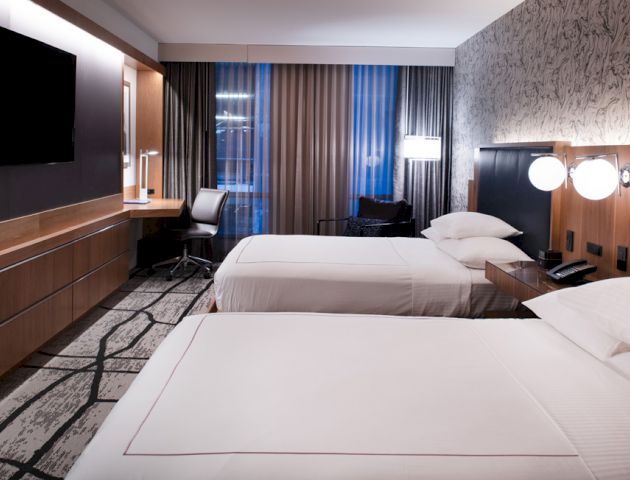 The image shows a hotel room with two neatly made beds, a flat-screen TV, a desk with a chair, and modern decor, featuring a patterned accent wall.