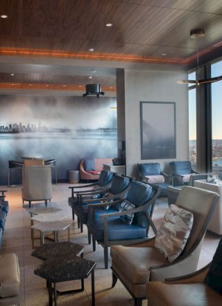 A modern lounge area with large windows, plenty of seating, and a panoramic view of a waterfront cityscape, including a Ferris wheel outside.