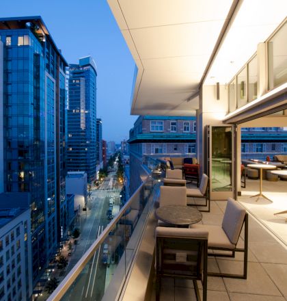 A modern cityscape featuring high-rise buildings at dusk, viewed from a spacious terrace with tables and chairs, partially enclosed by glass.