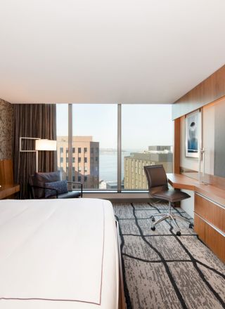 A modern hotel room with a large bed, TV, desk, and a view of buildings through a big window. The room features stylish decor and ample natural light.