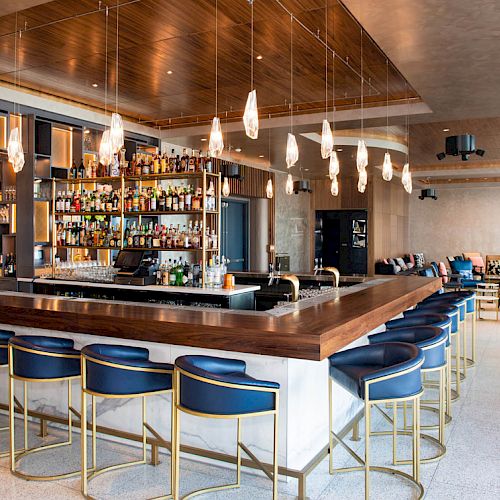 A modern bar with blue bar stools, a well-stocked liquor shelf, pendant lights, and a seating area in the background.