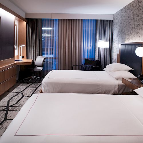 The image shows a modern hotel room with two neatly made twin beds, a wall-mounted TV, a desk with a chair, and stylish decor.