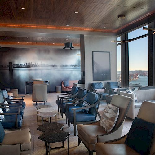 A lounge area with leather chairs, a mural of a cityscape, large windows with a water view, and modern decor in neutral tones is shown.