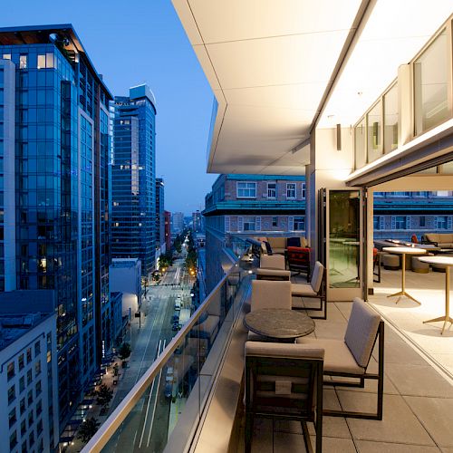 The image shows a modern cityscape from a high-rise balcony at dusk, with buildings and a street view. The balcony has outdoor seating.