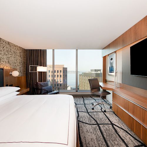 This image shows a modern hotel room with a large bed, a desk, a chair, a TV, and a view of buildings through a large window at the far end.