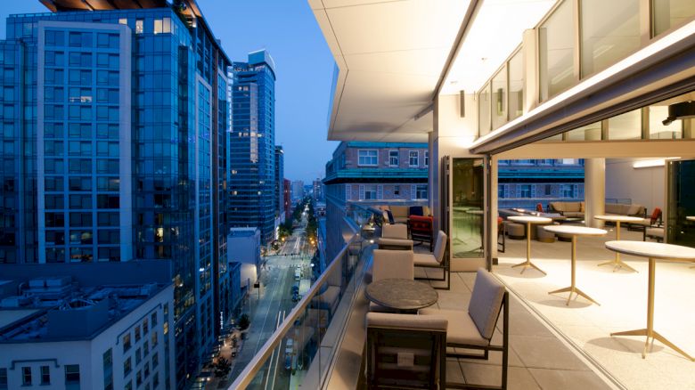 The image shows a modern urban balcony with seating and tables, overlooking a cityscape of tall buildings at dusk with street lights on.