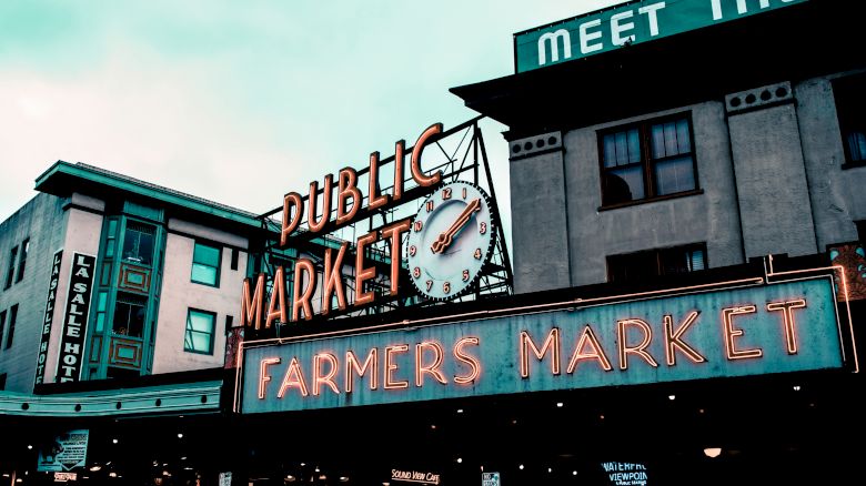 The image shows the iconic neon signs of a public farmers market, with surrounding buildings and a clock displaying a time close to noon or midnight.