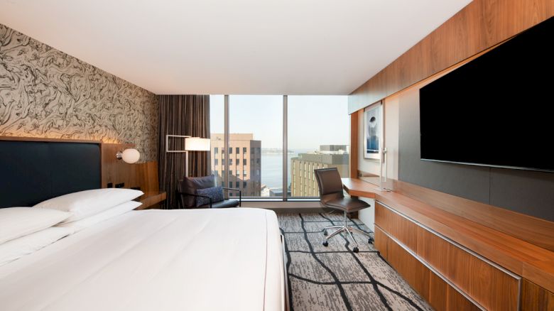 A modern hotel room featuring a large bed, a wall-mounted TV, a wooden desk, and a chair, with a window offering a city view.