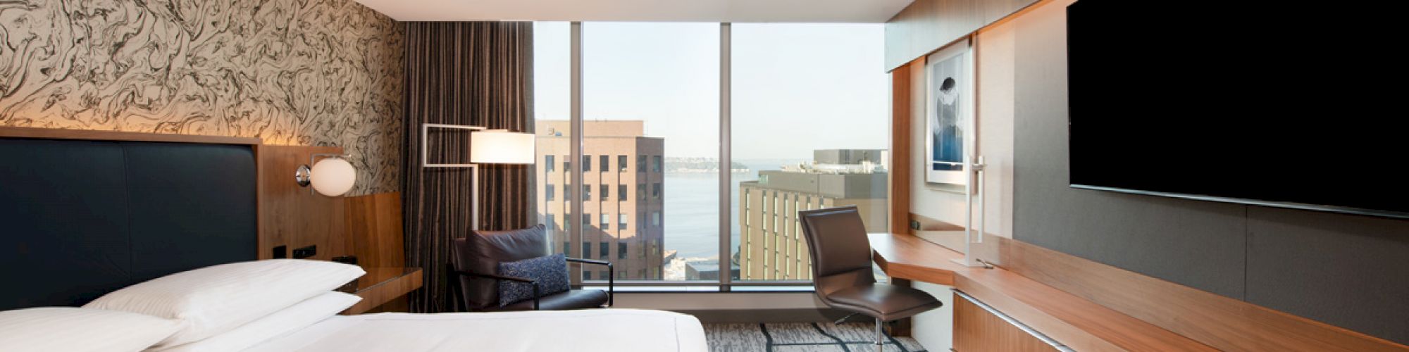 The image shows a modern hotel room with a large bed, wall-mounted TV, desk, chair, and cityscape view through a floor-to-ceiling window.