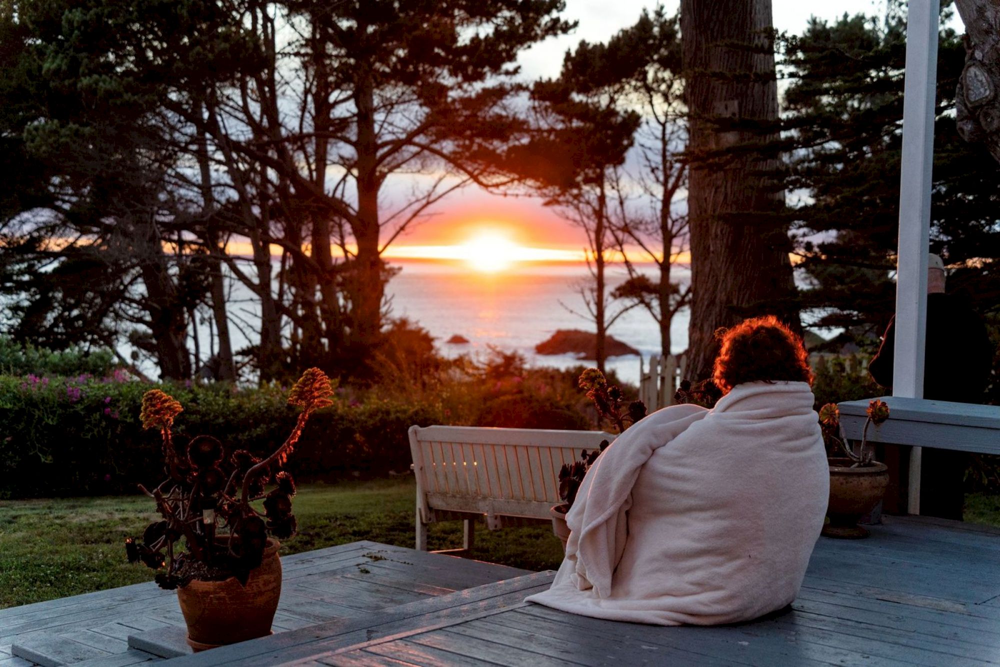 A person sits wrapped in a blanket on a deck, watching the sunset over a coastal landscape with trees and ocean visible in the background.