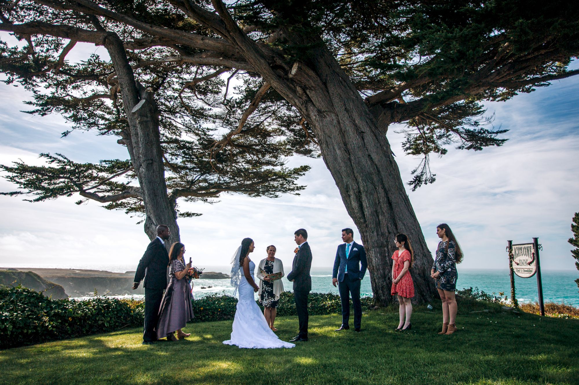 A wedding ceremony is taking place outdoors under large trees with an ocean view. The couple is surrounded by guests and an officiant.