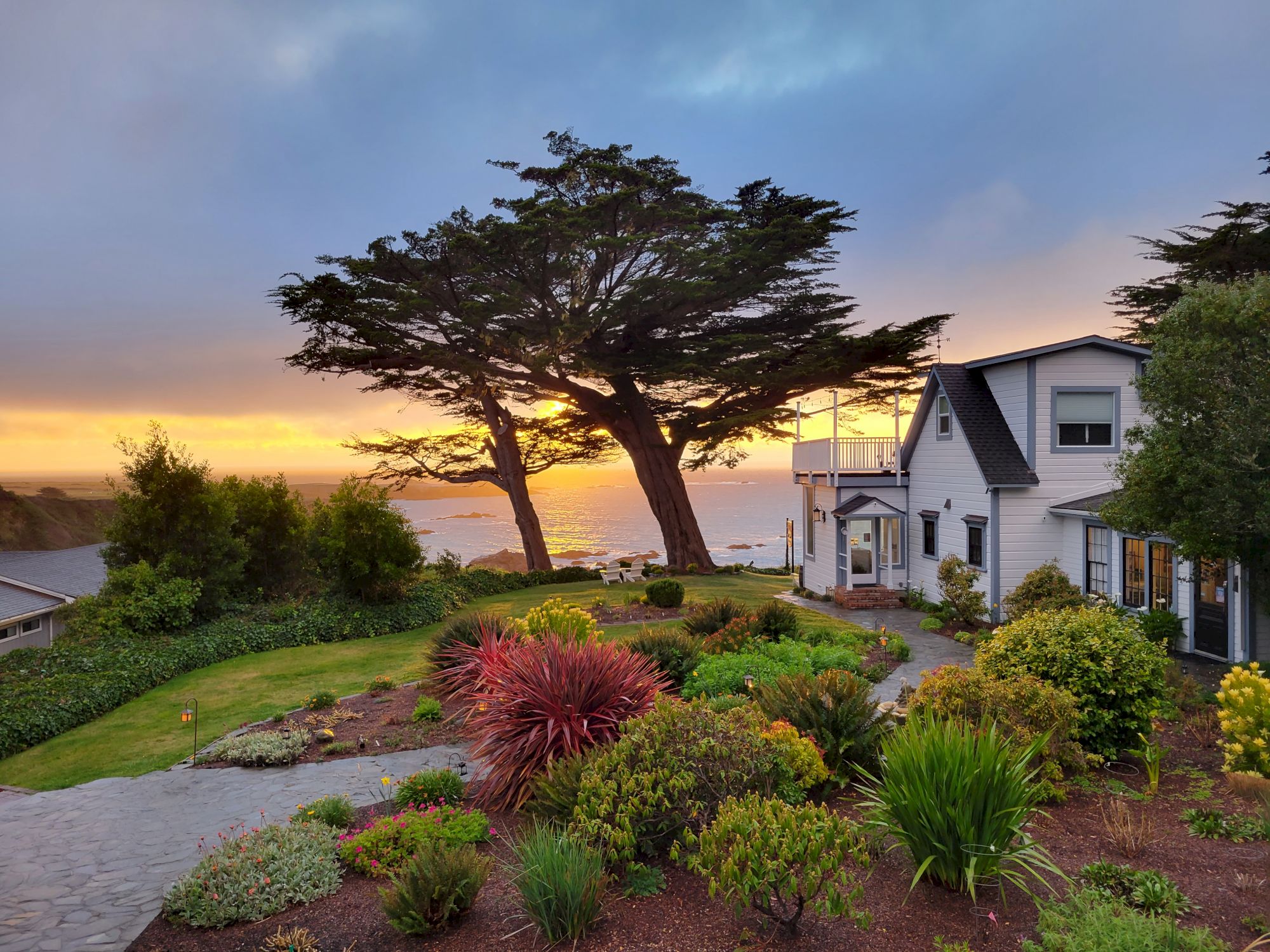 A scenic coastal view features a modern house, diverse garden plants, tall trees, and the ocean at sunset in the background.