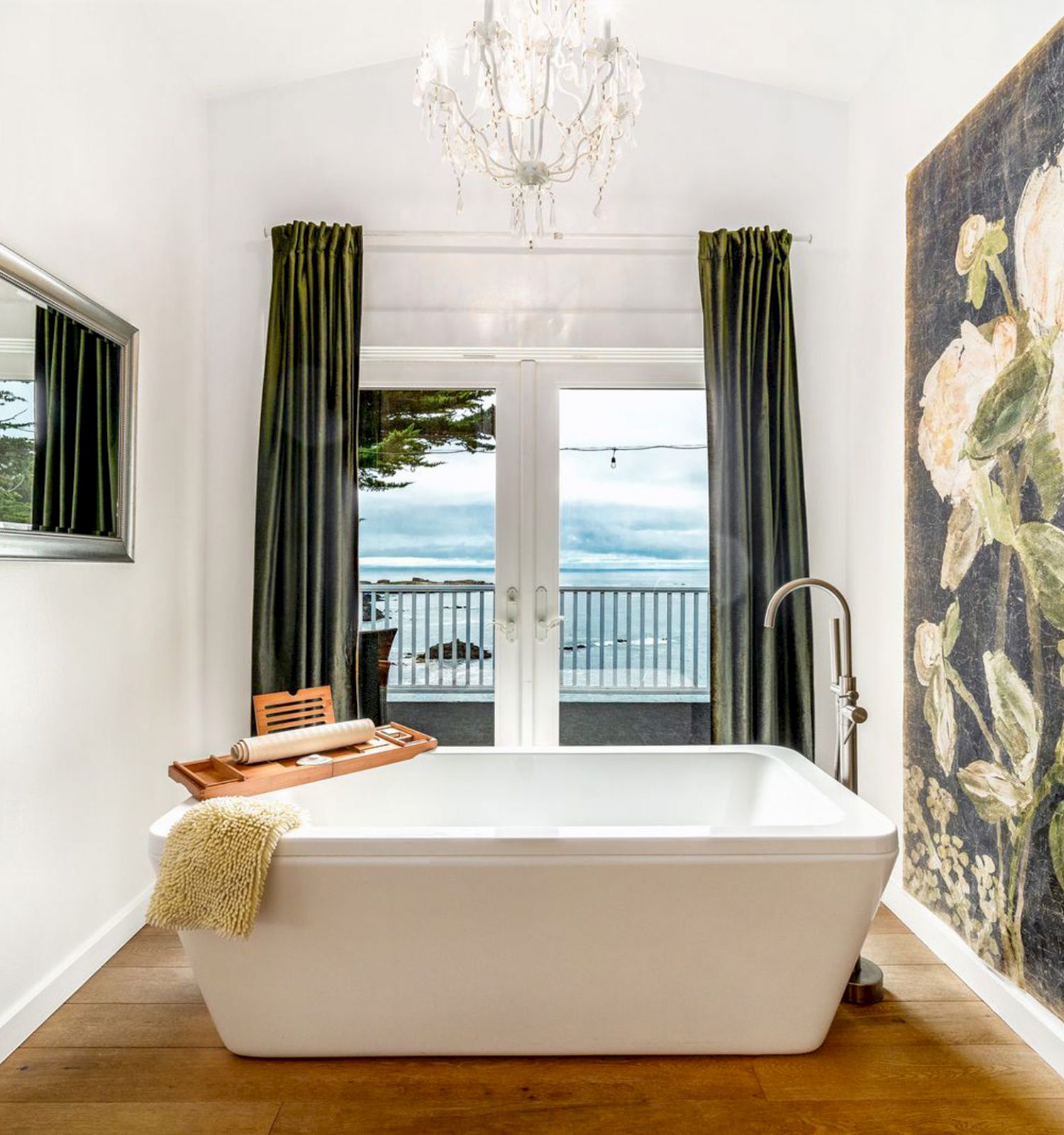 A modern bathroom with a white bathtub, large floral artwork, a chandelier, a mirror, and doors opening to a balcony with an ocean view.