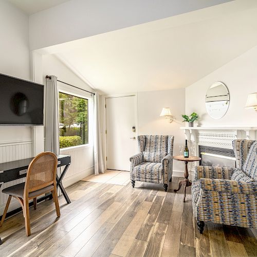 A cozy living space with two armchairs, a wall-mounted TV, a desk and chair, a window, and a mirror above a fireplace.