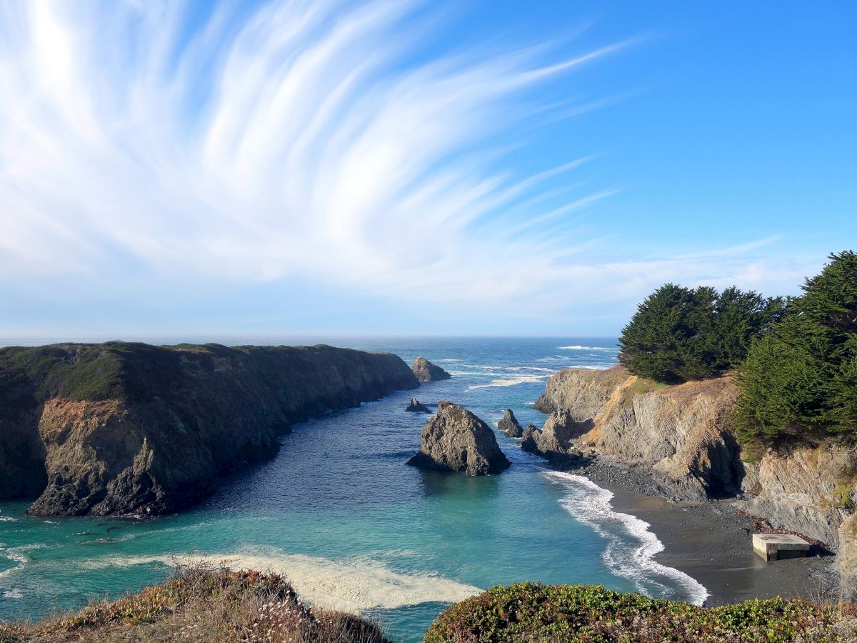 A scenic coastal landscape features rocky cliffs, turquoise waters, lush green trees, and a bright, expansive sky with wispy clouds ending the sentence.