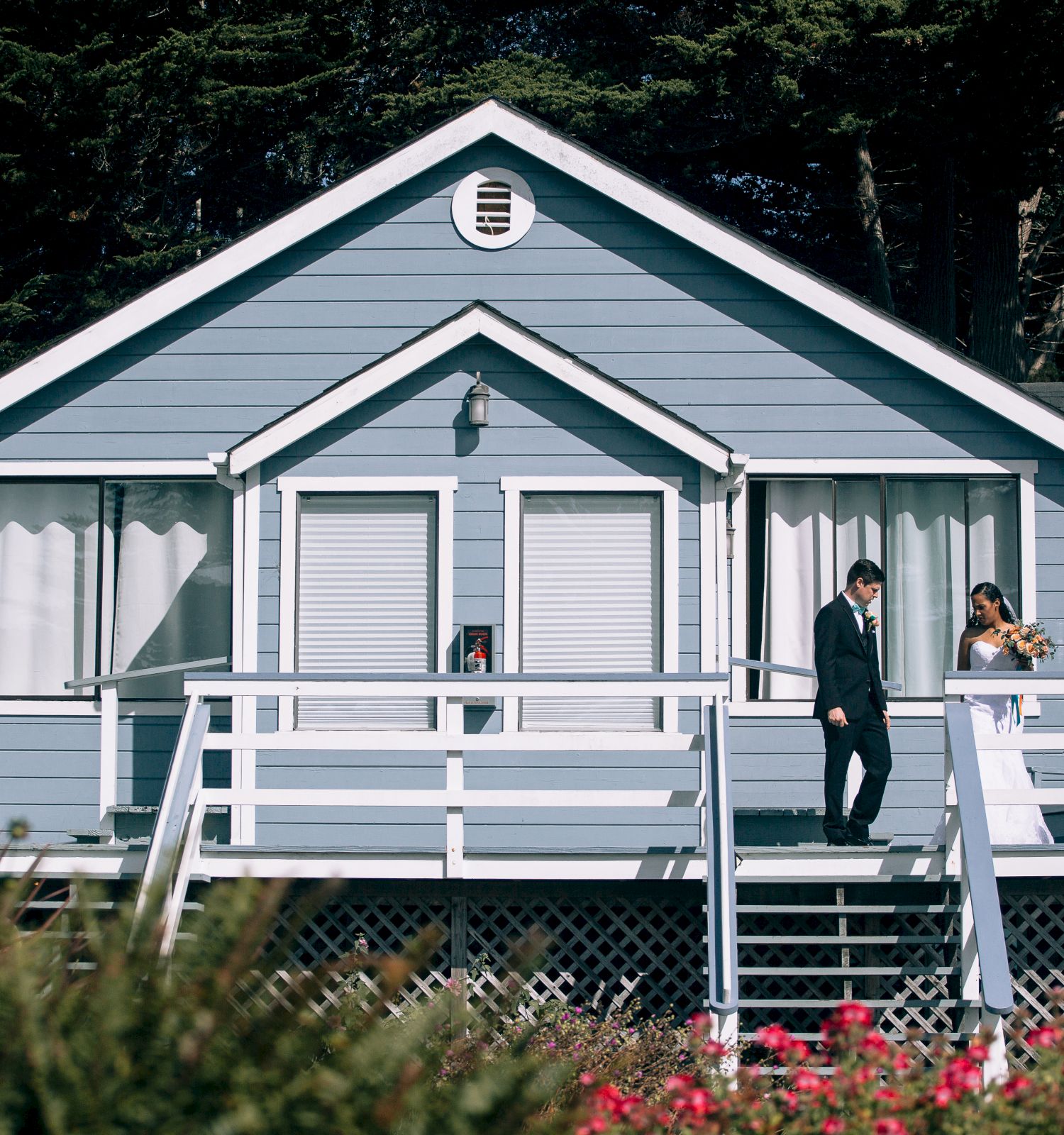 A blue house with a white railing, two people standing on the porch, surrounded by greenery and flowers, with trees in the background.