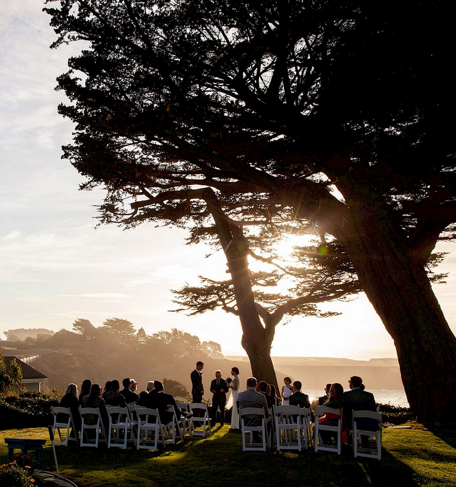 A group of people are gathered outdoors for an event or ceremony by a scenic coastline, with large trees and the sun setting in the background.