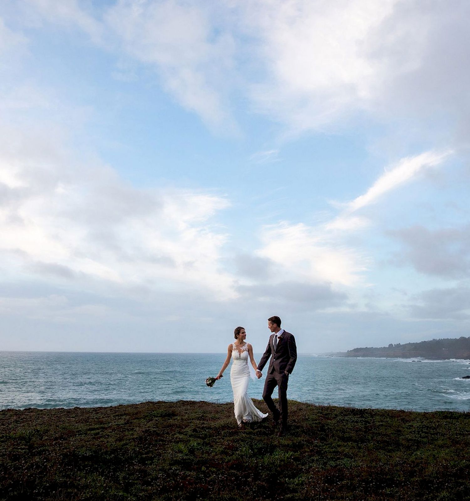 A couple in wedding attire walks along a cliff by the ocean with a dramatic sky and coastline in the background, holding hands.